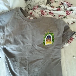 Grå t-shirt från Casablanca Paris - Snygg grå t-shirt från Casablanca Paris med färgglad tryckt grafik på både bröstet och ryggen. Motivet föreställer en byggnad i nattmiljö omgiven av gröna blad och texten 'Avenida Casablanca'. Klassisk passform och rund hals. Perfekt för dig som gillar unika prints.