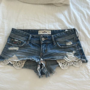 Jeansshorts med spets från Hollister - Snygga blå jeansshorts från Hollister med slitna detaljer, fransig kant och vit spets längs bensluten.  En låg midja med en snygg vardags vibbar. Super fina till stranden eller bara till någon fin tröja. Om det behövs mått eller beskrivningar meddela mig så fixar jag💕