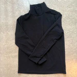 Svart tröja i bomull - En svart tröja med lång ärm och klassisk hög krage. Tröjan är i mjuk bomull och har en enkel, stilren design som funkar till allt. Perfekt att styla lager på lager under jackor eller skjortor.