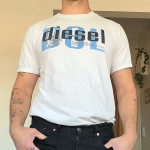 Vintage diesel T-shirt  - Vit t-shirt från Diesel med stort blått och svart logotryck på bröstet och mindre tryck i nacken. Klassisk passform med rund hals och korta ärmar. Skön och lätt i bomull, perfekt till jeans eller shorts. Sitter som en M