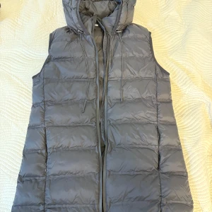 Svart lång dunväst från Zara - Lång, quiltad dunväst från Zara i svart med huva och dragsko. Västen har dragkedja framtill och två fickor med dragkedja på sidorna. 