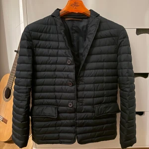 Dondup dunjacka  - Snygg marinblå quiltad dunjacka från Dondup med klassisk kavajkrage och tre knappar framtill. Jackan har två stora fickor med lock och är lätt vadderad för extra värme. Perfekt för dig som vill ha en stilren look med en twist. Passar både till jeans och chinos.