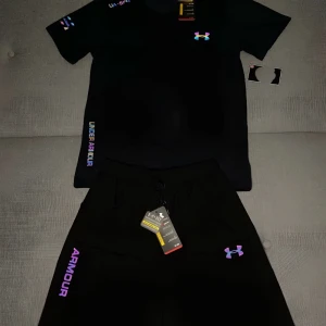 Svart Under Armour t-shirt med färgglada detaljer - Svart t-shirt från Under Armour med kort ärm och färgglada loggor och textdetaljer i regnbågsfärger. Tillverkad i ett lätt och sportigt material som andas, perfekt för träning. Snygg passform och modern look med UA-spec tryck på axeln.
