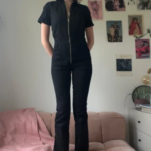 Svart jumpsuit med dragkedja från Mango - Snygg svart jumpsuit från Mango med korta ärmar, krage och hel dragkedja framtill. Modellen är figurnära med långa ben! Fickor både fram och bak.