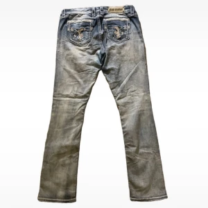 Rock Revival ljusblå jeans med broderi - Snygga ljusblå jeans från Rock Revival med slitningar och broderade bakfickor. Jeansen har raka ben, tydliga kontrastsömmar och coola detaljer på fickorna. Perfekt för dig som vill sticka ut med en unik denimstil. Skulle säga att dem är lite längre i benen också!💗 Lite slitna.