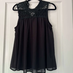 Svart ärmlös blus med spets från ONLY - Supersnygg svart blus från ONLY i storlek 36. Toppen är ärmlös med ett flowy fall och har en detaljerad virkad spets upptill samt knäppning i nacken. Perfekt när du vill ha något stilrent men ändå med coola detaljer.