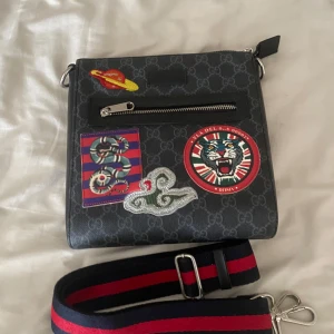 GG axelväska med patches - Snygg svart axelväska från guci  med GG-monogram och coola patches framtill, bland annat ormar och tiger. Väskan har en bred, randig axelrem i rött och marinblått samt dragkedja och ytterfack. Materialet är skinn och detaljerna är i silverfärgad metall.