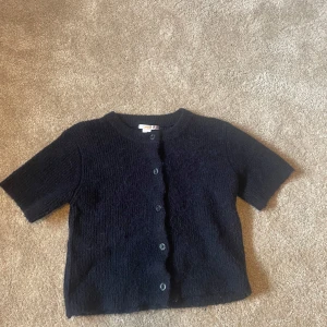 Svart stickad kofta från Gina Tricot - Kortärmad svart stickad kofta från Gina Tricot med rund halsringning och knappar framtill. Koftan har en mjuk och mysig känsla, perfekt att slänga över en topp eller klänning. Enkel och stilren design som funkar till många outfits.