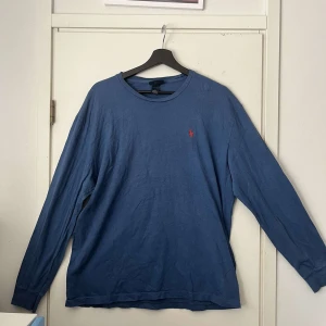 Blå långärmad tröja Polo Ralph Lauren - Snygg blå långärmad tröja från Polo Ralph Lauren i 100% bomull. Klassisk rund hals och ikonisk röd broderad logga på bröstet. Perfekt för dig som gillar stilren och enkel design med en touch av preppy vibe.