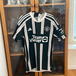 Manchester United bortaställ 2023-2024 Size S - Manchester Uniteds officiella bortatröja från Adidas i storlek S. Tröjan är randig i mörkgrönt, vitt och rött med klubbmärke och TeamViewer-logga på bröstet. Klassisk V-ringning, korta ärmar och tillverkad i lätt, ventilerande polyester. Har själv en stor fotbollströjssamling så kan garantera att varan till 100% är äkta!