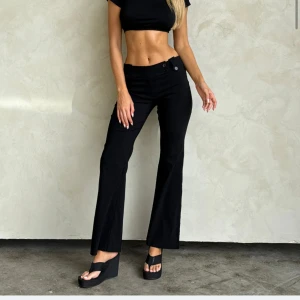 Svarta bootcut byxor med fickdetaljer - Snygga svarta bootcut byxor med låg midja och coola fickdetaljer bak. Byxorna har en klassisk passform med utsvängda ben och knappar både fram och bak. Perfekta för dig som gillar en stilren och trendig look.