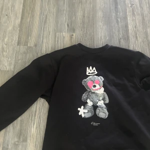 Svart sweatshirt Closure LDN med nalletryck - Svart sweatshirt från Closure LDN i storlek S med ett stort tryck av en grå nalle med rosa detaljer och en vit krona på bröstet. Tröjan har rund hals och långa ärmar, perfekt för dig som gillar streetstyle och coola prints.