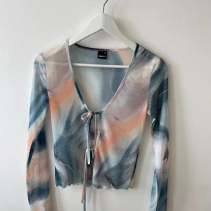Mesh topp med knyt från Gina Tricot - Säljer en mesh topp från Gina Tricot i storlek XS. Toppen har ett abstrakt mönster i blått, vitt, orange och grått, v-ringning och knyt framtill. Långärmad och croppad modell med vågig kant nertill. Perfekt för dig som gillar trendiga detaljer och färg.