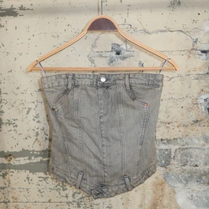 Grå jeanskorsett med dragkedja - Cool grå korsettopp i jeansmaterial med markerade sömmar och bälteshällor. Framsidan har knapp- och fickdetaljer, medan baksidan har smock och dragkedja för en tight passform. Perfekt för dig som vill sticka ut med en edgy denimlook.
