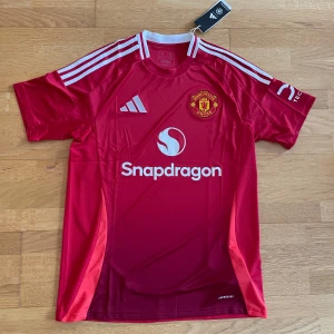 Manchester United matchtröja Adidas M - Manchester Uniteds officiella matchtröja från Adidas i rött med vita detaljer och klubbmärke broderat på bröstet. Tröjan har korta ärmar, vit krage och tre vita ränder på axlarna. Tillverkad i lätt och ventilerande polyester med Snapdragon sponsortryck.