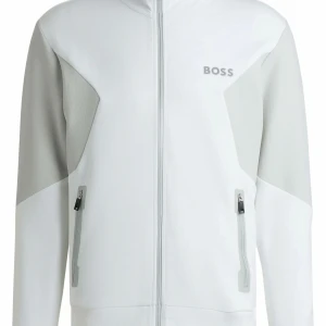 Vit BOSS tröja  - Snygg vit track jacket från BOSS med grå paneler på ärmar och axlar. Jackan har hel dragkedja framtill, två fickor med dragkedja och diskret BOSS-logga på bröstet. Perfekt för en clean och sportig look. Mycket bra skick utan tecken på användning.