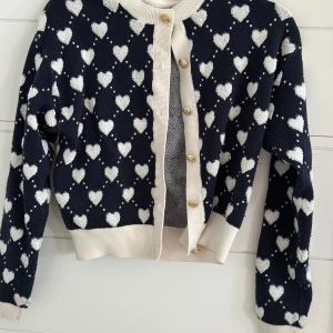 Mörkblå kofta med hjärtan Holly & white - Supersöt mörkblå kofta från Holly & Whyte med vita hjärtan över hela tröjan. Krämvita ribbade muddar och knappslå med guldiga knappar. 