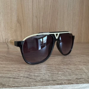 Louis Vuitton solglasögon - Snygga solglasögon från Louis Vuitton med svart båge och guldiga detaljer. Modellen har en klassisk pilotform och mörka linser. Sidorna är märkta med Louis Vuitton-loggan och bågarna har en lyxig känsla med guldaccenter.