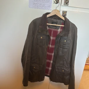 Brun parkajacka - Säljer en brun parkajacka från Lindex med snyggt rutigt foder i rött och vitt. Jackan har flera fickor med knappar, hög krage i manchester och dragkedja framtill. Perfekt för dig som gillar en klassisk och avslappnad stil. 