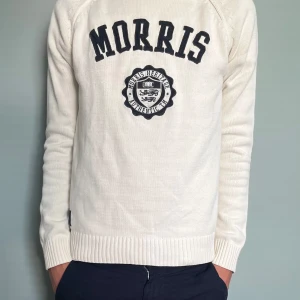 Vit stickad tröja från Morris - Vit stickad tröja från Morris med broderad logga och text på bröstet. Tröjan har svarta armbågslappar, ribbade muddar och rund hals. Perfekt för dig som gillar klassisk och sportig stil.