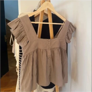 Beige blus med volangärmar - Supersöt beige blus med breda axelband och volangdetaljer på ärmarna. Knappar framtill och luftig passform gör den perfekt för varma dagar. Tillverkad i ett mjukt bomullstyg som känns skönt mot huden.