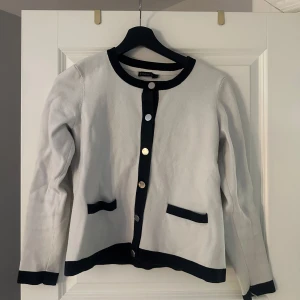 Vit kofta med svarta detaljer Stockh LM - Snygg vit kofta från Stockh LM med svarta kanter och silverfärgade knappar. Långärmad modell med två dekorativa fickor framtill. Perfekt för dig som gillar en klassisk men trendig look.