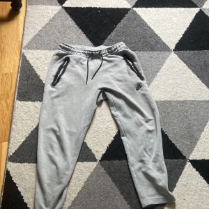Gråa Nike teck byxor - Gråa mjukisbyxor från Nike i storlek M. Byxorna har snörning i midjan, dragkedjor på fickorna och en klassisk Nike-logga på ena benet. Perfekta för chill eller träning, tillverkade i mjukt bomullsmaterial.