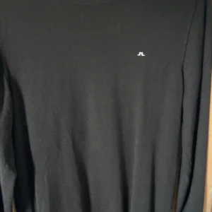 Svart långärmad tröja från J.Lindeberg - Snygg svart långärmad tröja från J.Lindeberg med diskret logga på bröstet. Enkel och stilren design som passar perfekt till jeans eller chinos. Mjuk och skön känsla, perfekt för lager-på-lager under kyligare dagar.