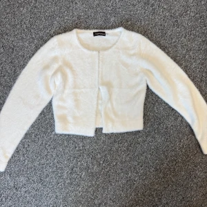 Vit fluffig cardigan jacka från Alessa - Supermjuk och fluffig vit cardigan jacka från Alessa. Croppad modell med lång ärm och rund halsringning. Stängs med små hakar framtill. Perfekt för dig som vill ha en mysig och stilren look med lite extra fluff.