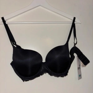Svart push-up bh från Dorina - Snygg svart push-up bh från Dorina med tunna axelband och spetsdetaljer längs nederkanten. Bh:n har vadderade kupor och klassisk knäppning bak. Perfekt för dig som vill ha en stilren och bekväm bh med lite extra lyft.