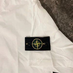 Vit Stone Island t-shirt med patch - Vit t-shirt från Stone Island med klassisk rund hals och korta ärmar. På vänster ärm sitter den ikoniska svarta patchen med gult och grönt kompassmärke. T-shirten är i mjuk bomull och har en clean, stilren look som passar perfekt till jeans eller shorts.