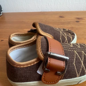 Bruna sneakers med spänne och mönster - Unika bruna sneakers i textil med snygga broderade mönster och kontrasterande ljusbruna kanter. Skorna har breda remmar i konstläder med metallspänne och vit platt sula. Perfekta för dig som gillar en cool och annorlunda stil.