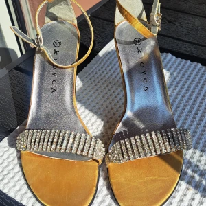 Guldiga pumps med strass från Joyca - Glittriga pumps i guldigt satin från Joyca med smal klack och öppen tå. Rem runt vristen med spänne och bred rem framtill dekorerad med strass. Perfekta för dig som vill sticka ut med en lyxig och festlig look.