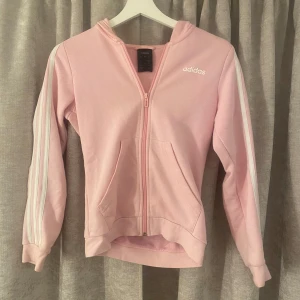 Rosa Adidas zip hoodie - Snygg ljusrosa zip hoodie från Adidas med klassiska vita ränder längs ärmarna och vit logga på bröstet. Jackan har huva, dragkedja framtill och två fickor. Perfekt för en sportig och chill stil.