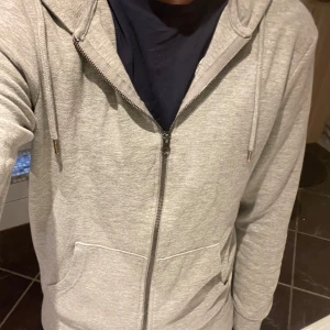 Grå zip-hoodie från Jack & Jones S - Säljer en grå hoodie från Jack & Jones Essentials i storlek S. Den har dragkedja framtill, huva med snören och två fickor. Perfekt för chill dagar eller när du vill ha en enkel och clean look. Materialet är mjukt och skönt bomullstyg.