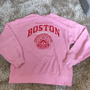 Rosa Boston sweatshirt från Divided - Säljer en rosa sweatshirt från Divided med tryck i vitt och rött där det står 'Boston Massachusetts'. Tröjan har rund hals, långa ärmar och ribbade muddar. Perfekt för dig som gillar college-stil och vill ha en skön och snygg tröja till vardags.