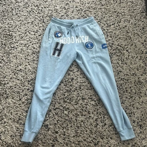 Ljusblå mjukisbyxor från Hoodrich - Snygga ljusblå mjukisbyxor från Hoodrich med broderade patches och tryck framtill. Byxorna har snörning i midjan, muddar vid bensluten och två sidofickor. Perfekta för en chill och avslappnad stil.