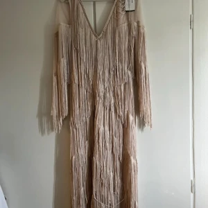 Beige långklänning med fransar - Supercool beige långklänning med massor av fransar och v-ringning. Klänningen har långa ärmar och ger en riktigt bohemisk och unik vibe. Perfekt för dig som vill sticka ut och älskar detaljer som rör sig när du går.