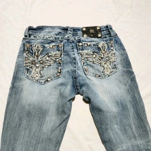 Slim bootcut jeans med broderi, stl 28 - Ljusblå jeans från Miss Me i bootcut-modell med slim passform. Snygga broderade bakfickor med nitar och korsdetaljer. Jeansen har slitningar och en rivning på ena knät för en edgy look. Klassisk femficksmodell och grova kontrastsömmar.