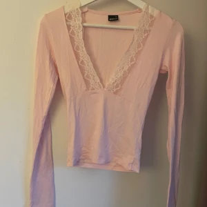 Ljusrosa långärmad topp Gina Tricot - Supersöt ljusrosa långärmad topp från Gina Tricot med djup v-ringning och spetsdetaljer längs urringningen. Toppens material är mjukt och stretchigt, perfekt för en feminin och trendig look. Passar dig som gillar romantiska detaljer och en smickrande passform.