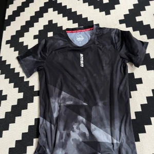 Svart camo t-shirt från USWE - Säljer en svart t-shirt från USWE i storlek S. T-shirten har ett diskret camouflagemönster i olika grå toner och är tillverkad i ett lätt och snabbtorkande syntetmaterial. Perfekt för träning eller aktiva dagar. Klassisk passform med korta ärmar och rund hals.