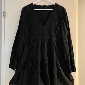 Svart kort klänning från Zara - Supersnygg svart klänning från Zara med v-ringning och långa ärmar. Klänningen har en luftig, volangig kjol och fina broderade detaljer framtill. Perfekt för dig som gillar en enkel men chic look.