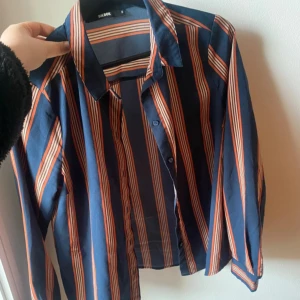 Randig skjorta från Bik Bok, stl S - Snygg skjorta från Bik Bok i storlek S med blå bas och orangea ränder. Skjortan har klassisk krage, knäppning framtill och långa ärmar. Materialet känns lätt och luftigt, perfekt för dig som gillar färg och mönster. Passar till jeans eller kjol för en cool look.