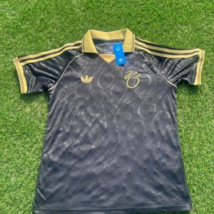 Jude Bellingham Adidas fotbollströja L - Snygg svart fotbollströja från Adidas i storlek L med guldiga detaljer och broderad logga. Tröjan har korta ärmar, klassisk krage och diskret mönster i tyget. Guldiga ränder på axlarna och kragen ger en exklusiv känsla. Tillverkad i lätt och ventilerande material.