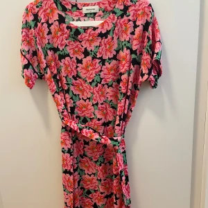 Blommig klänning från Modström - Färgstark klänning från Modström med stora rosa och röda blommor på svart botten. Modellen har korta ärmar, knytband i midjan och en avslappnad passform. Perfekt för dig som vill sticka ut med ett coolt blommönster.