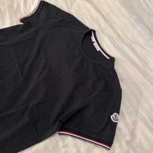 Svart Moncler t-shirt med logga - Svart t-shirt från Moncler i slim fit-modell. Klassisk rund halsringning och korta ärmar med röd, vit och blå rand med Moncler-logga på vänster ärm. Skön bomullskvalitet och stilren design som sticker ut med snygga detaljer. Denna tshirt har aldrig använts så den är i nyskick och har fortfarande taggarna på. Pris kan diskuteras! och kan mötas upp i Skåne. 