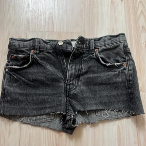 Svarta jeansshorts från Zara - Snygga svarta jeansshorts från Zara med råa, fransiga kanter och klassisk femficksdesign. Shortsen har dragkedja och knapp framtill samt slitna detaljer för en cool, avslappnad look. Perfekta till sommaren och lätta att matcha med allt. Högmidjade och korta där bak!!