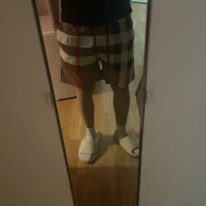 Rutig Burberry shorts i beige och brunt - Säljer ett par klassiska Burberry shorts i rutigt mönster med beige, bruna, svarta och vita toner samt en röd detalj. De har elastisk midja och meshfoder, perfekt för sommaren. Ikonisk design som sticker ut och känns riktigt lyxig. Kvitto finns!