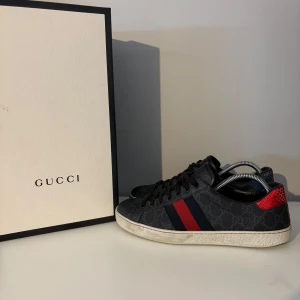 Gucci sneakers med röd och blå detalj - Snygga Gucci sneakers i svart med klassiskt GG-mönster, röda och blå ränder på sidan samt metallicröd och metallicblå häl. Skorna har snörning och platt sula. Materialmix av skinn och syntet, perfekt för dig som vill sticka ut med lyxig streetstyle.