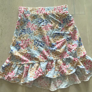 Blommig volangkjol från Gina Tricot - Supersöt kort kjol från Gina Tricot i blommigt mönster med volangkant. Kjolen har en mix av ljusa färger som rosa, blå, beige och vit, och är perfekt för en somrig look.